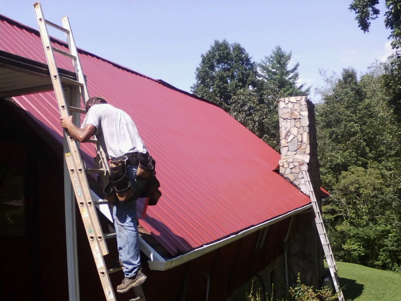 Roofer on a ladder beside a red metal roof and stone chimney — Soffit & Fascia Repair by Roofing Brothers in Paragonah